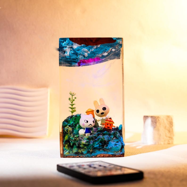 Może przedstawiać: Prostokątny element artystyczny z żywicy z miniaturową dioramą. Przedstawia postacie z kresk&oacute;wek, w tym białego kota i kr&oacute;lika, na niebiesko-zielonej podstawie. G&oacute;rna część ma abstrakcyjną niebieską i brązową sztukę.