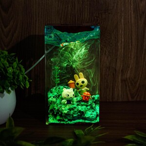 Może przedstawiać: Prostokątna żywiczna lampka nocna z zielonym, świecącym wnętrzem. W środku znajdują się małe figurki kota, kr&oacute;lika i czerwonej latarni. Tło to ciemna drewniana powierzchnia.