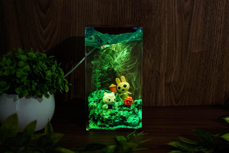 Może przedstawiać: Prostokątna żywiczna lampka nocna z zielonym, świecącym wnętrzem. W środku znajdują się małe figurki kota, kr&oacute;lika i czerwonej latarni. Tło to ciemna drewniana powierzchnia.