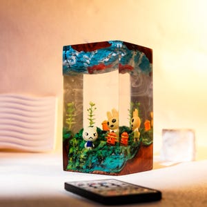 Może przedstawiać: Prostokątny element artystyczny z żywicy z miniaturową dioramą wewnątrz, przedstawiający postacie z kresk&oacute;wek, rośliny i małe konstrukcje. Na g&oacute;rze znajduje się niebiesko-czerwony abstrakcyjny wz&oacute;r. Pilot znajduje się na pierwszym planie.