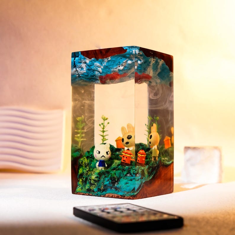 Może przedstawiać: Prostokątny element artystyczny z żywicy z miniaturową dioramą wewnątrz, przedstawiający postacie z kresk&oacute;wek, rośliny i małe konstrukcje. Na g&oacute;rze znajduje się niebiesko-czerwony abstrakcyjny wz&oacute;r. Pilot znajduje się na pierwszym planie.