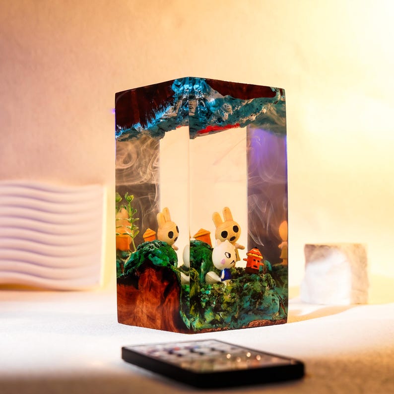 Może przedstawiać: Prostokątna lampa z żywicy z leśną scenerią z małymi figurkami kr&oacute;lik&oacute;w. Lampa ma drewnianą podstawę i niebiesko-czerwony wierzch. Scena obejmuje zielone liście i małe pomarańczowe konstrukcje. Pilot znajduje się na pierwszym planie.