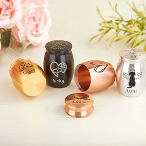 Puede incluir: Una colección de pequeñas urnas para mascotas en varios colores, incluyendo dorado, cobre, negro y plateado. Algunas urnas presentan alas de ángel, huellas de patas y las palabras "Sunny", "lucky" y "Aunt".