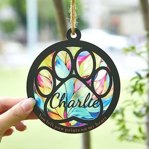 May include: A round, black pet memorial ornament with a paw print design. The ornament features the name "Charlie" and the text "You left paw prints on my heart." The paw print is filled with colorful leaves, and it hangs from a twine string.