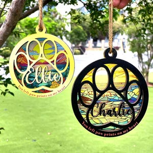 Op de afbeelding: Twee ronde, gelaagde houten ornamenten met pootafdrukontwerpen. Eén is geel met "Ellie" en de tekst "You left paw prints on my heart." De andere is zwart met "Charlie" en dezelfde tekst. Beide hebben een kleurrijke achtergrond in glas-in-loodstijl.