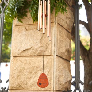 May include: Copper-colored wind chimes hanging in front of a stone wall. The chimes have several long tubes and a wooden wind catcher with engraved text and a heart symbol. The chimes are suspended by a black string.