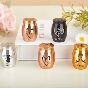 May include: A selection of small pet urns in silver, copper, and gold. Each urn has a heart, paw print, or angel wing design, with names such as "Enzo" and "Sunny". The urns are displayed on a white surface. Measurements are in centimeters.