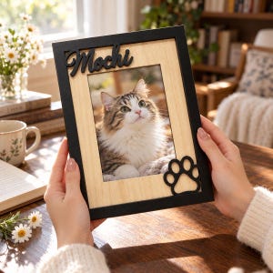 May include: A rectangular picture frame with a black border and a light wood-toned inner frame. The top features the name Mochi in black script, and the bottom right corner has a black paw print cutout. A photo of a fluffy cat is inside.