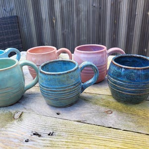 Op de afbeelding: Een verzameling handgemaakte keramische mokken in verschillende kleuren, waaronder turquoise, roze en blauw. Elke mok heeft een afgeronde vorm met een handvat en horizontale getextureerde lijnen. De mokken staan op een verweerd houten oppervlak.