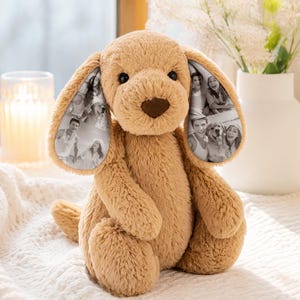 Puede incluir: Un juguete de peluche con forma de perro de color beige, con pelaje suave y texturizado y una nariz marrón oscuro. El interior de cada oreja caída presenta una impresión fotográfica personalizada en blanco y negro.