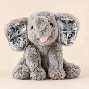 Puede incluir: Un juguete de peluche de elefante gris con una textura suave y esponjosa y una pequeña lengua rosa. El elefante presenta orejas grandes y caídas, cada una con un collage de fotografías en blanco y negro impresas en la tela de las orejas.