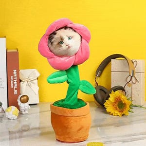 Op de afbeelding: Een pluche speeltje met een kattengezicht in het midden van een roze bloem, bevestigd aan een groene stengel met twee bladeren, alles in een bruine stoffen pot. De kat heeft witte, bruine en zwarte vacht met groene ogen en een zachte textuur.