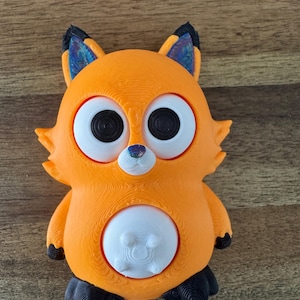 Könnte beinhalten: Eine 3D-gedruckte orangefarbene Fuchsstatuette mit großen, runden Augen und einem weißen Bauch. Der Fuchs hat schwarze Füße und Ohren mit schillernden blau-violetten Akzenten. Die Figur ist mit einer strukturierten Oberfläche versehen.