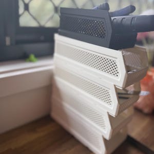 May include: A stack of four white plastic storage bins with a perforated design. A black plastic item with a mesh front sits on top. The bins are on a wooden surface, near a window.