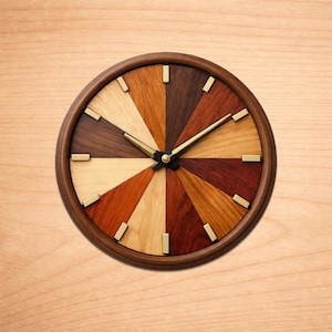 May include: A round wooden clock with a dark brown frame and a face divided into segments of various wood tones. Gold-colored rectangular markers indicate the hours. The clock hands are gold, and the clock is set against a light wood background.