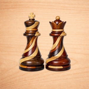 May include: Two wooden chess pieces: a king and a queen. The king has a cross on top, and the queen has a crown. Both pieces are made of dark and light wood, with a striped design. The pieces are on a light brown wooden surface.