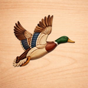 Puede incluir: Escultura de madera de un pato en vuelo, con las alas extendidas. El pato presenta detalles en marrón, azul, verde y crema. La talla se encuentra sobre un fondo claro de vetas de madera, resaltando la forma del pato.