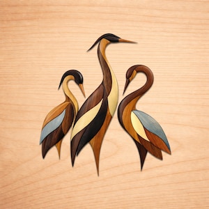 May include: Three wooden bird sculptures with long necks and pointed beaks. The birds are made of various wood tones, including brown, black, and tan, with accents of blue and gold. The birds are arranged on a light wood background.