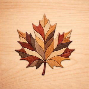 May include: A decorative maple leaf wall art piece crafted from multiple segments of wood in varying natural tones including light tan, golden yellow, deep reddish-brown, and dark chocolate brown, arranged in a symmetrical marquetry pattern.