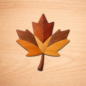 Puede incluir: Una hoja de arce de madera con un diseño de mosaico detallado. La hoja está compuesta por varios tonos de madera marrón, desde beige claro hasta caoba oscuro. El tallo es de color marrón oscuro, sobre un fondo claro de vetas de madera.