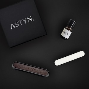 Könnte beinhalten: Eine schwarze Box mit dem Wort "ASTYN." darauf gedruckt, eine kleine braune Glasflasche mit schwarzem Verschluss und dem Wort "ASTYN" auf dem Etikett, ein weißes rechteckiges Objekt und ein schwarzer und brauner Holz-Lufterfrischer für das Auto mit dem Wort "ASTYN" darauf gedruckt.