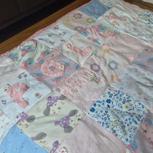 May include: A pastel-coloured patchwork quilt with squares of floral, teddy bear, and letter designs. The quilt features the embroidered word "Mummy" and is edged with pink fabric. The overall design is soft and inviting, suitable for a nursery.