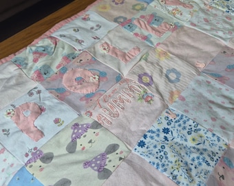 Puede incluir: Un edredón de retazos en colores pastel, con cuadrados con diseños florales, de osos de peluche y letras. El edredón incluye la palabra bordada "Mummy" y está bordeado con tela rosa. El diseño general es suave y acogedor.