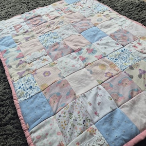 May include: A handmade patchwork quilt with a pink border. The quilt is composed of pastel-coloured fabric squares, including light blue, pink, and white. Some squares feature floral and animal prints. The quilt has a soft, cosy appearance and is likely handmade.