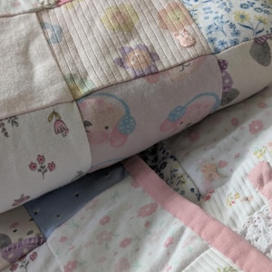 May include: A patchwork quilt with a variety of fabric patterns and colours. The quilt includes floral prints, teddy bear motifs, and ribbed fabric sections. Soft pinks, blues, and whites dominate the colour palette, with decorative buttons adding detail. The quilt appears to be handmade.