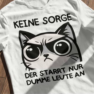 Könnte beinhalten: Weißes T-Shirt mit einem grimmigen Katzendesign und dem deutschen Text: "KEINE SORGE DER STARRT NUR DUMME LEUTE AN". Die Katze hat einen finsteren Blick und große Augen. Text und Katze sind in Schwarz und Weiß.