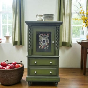 May include: A green cabinet with a glass door displaying a floral design. The cabinet has two drawers with white knobs and rope-style trim. A basket of red apples is placed nearby.
