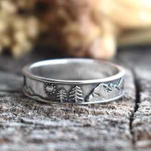 May include: A silver band ring featuring a mountain, sun, and pine tree design.