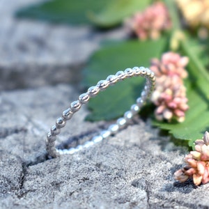 May include: A silver beaded ring with a simple design. The ring is lying on a textured surface with pink flowers.