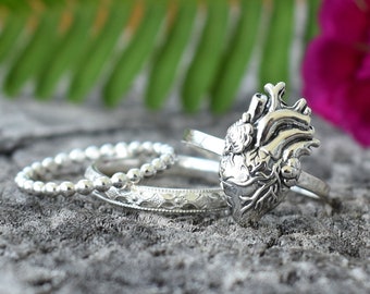 May include: A set of four silver rings, one featuring a detailed anatomical heart design. The rings are stacked on a gray textured surface.