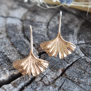 Könnte beinhalten: Ein Paar goldfarbene Ginkgo-Blatt-Ohrringe. Die Ohrringe sind zart und weisen ein detailliertes Design eines Ginkgo-Blattes auf.