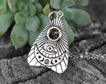 Könnte beinhalten: Eine silberne Anhängerkette mit einem stilisierten Augendesign in einem Sonnenstrahlenmuster. Der Anhänger hat ein detailliertes, graviertes Design mit Mond- und Punktakzenten. Die Halskette hat eine zarte, perlenbesetzte Kette, und der Anhänger wird vor einem natürlichen Holzgrund gezeigt.
