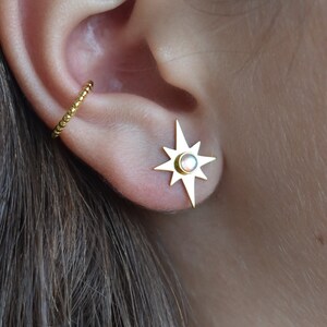 May include: Gold star-shaped earring with a white opal stone in the centre. The earring is worn in the upper lobe of the ear. A gold hoop earring is worn in the cartilage of the ear.