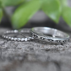 May include: Three silver rings stacked on a gray textured surface. The top ring has a decorative pattern of small circles and triangles. The middle ring has a beaded design. The bottom ring is plain.