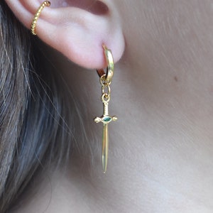 May include: Gold hoop earrings with a dangling sword charm. The sword has a green gemstone on the pommel.
