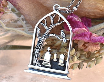 May include: Silver pendant necklace with a gothic graveyard design. The pendant depicts a tree with a bird, tombstones, and a cross inside an arched frame. The necklace has a fine chain.