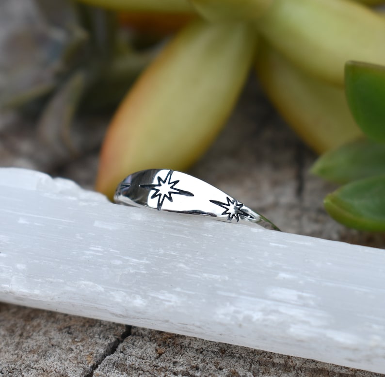 May include: A silver ring with two star designs. The ring is on a white surface.