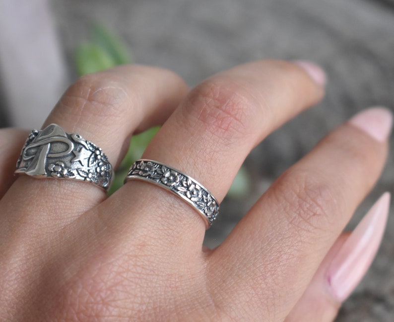 May include: Two silver rings with floral designs. The ring on the left has a cross and star design. The ring on the right is a thin band with a floral pattern.