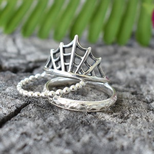 以下が含まれることがあります： 3つの銀のリングが積み重ねられています。一番上のリングは、繊細で透かし彫りのデザインで、クモの巣に似ています。真ん中のリングはビーズのバンドです。一番下のリングは、シンプルなテクスチャのバンドです。