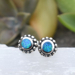 May include: A pair of silver stud earrings with a blue opal gemstone in each. The earrings have a beaded border around the opal.