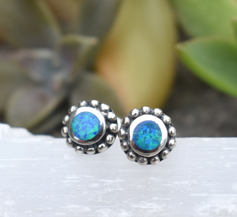 May include: A pair of silver stud earrings with a blue opal gemstone in each. The earrings have a beaded border around the opal.