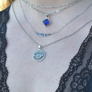 May include: A silver necklace with four chains and two pendants. The top pendant is a square with a dark blue gemstone. The bottom pendant is a round design with a sun and moon.