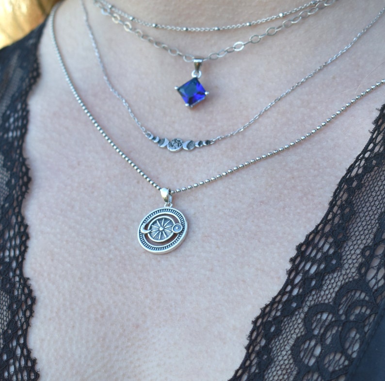 May include: A silver necklace with four chains and two pendants. The top pendant is a square with a dark blue gemstone. The bottom pendant is a round design with a sun and moon.