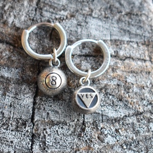 May include: A pair of silver hoop earrings with charms. One charm is a silver 8-ball and the other charm is a silver triangle with the word "YES" inscribed.