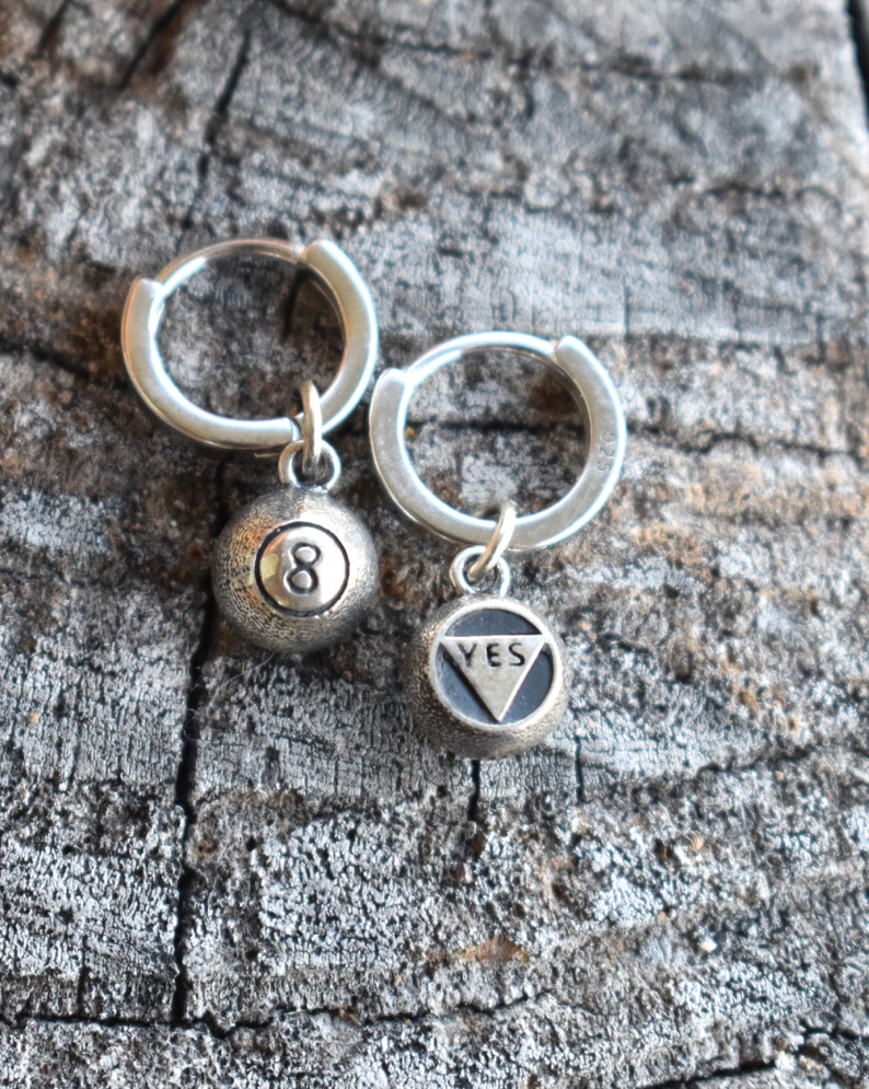 May include: A pair of silver hoop earrings with charms. One charm is a silver 8-ball and the other charm is a silver triangle with the word "YES" inscribed.