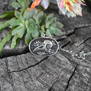 May include: A silver pendant necklace featuring a detailed, three-dimensional design of a crow skull with floral accents. The pendant is oval-shaped and hangs from a silver chain.